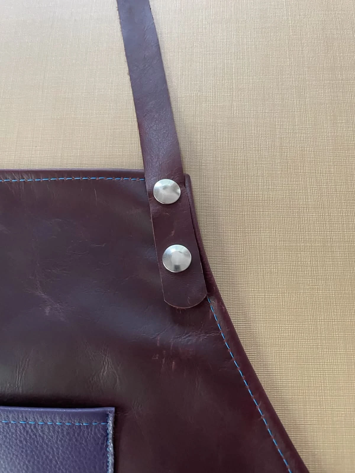 Two silver-tone fasteners on a burgundy leather neck strap beside blue contrast stitching