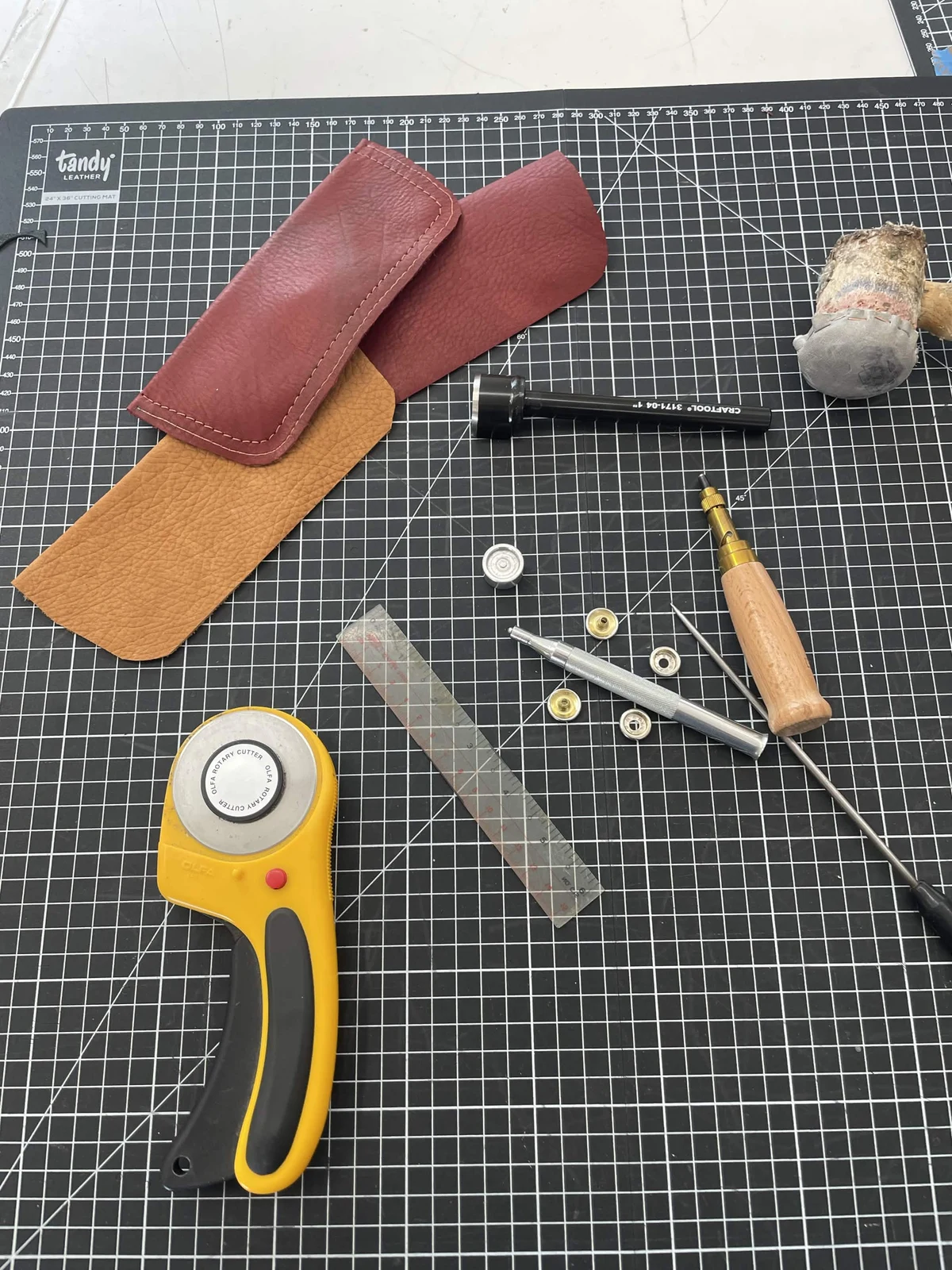 Leather pocket pieces, rotary cutter, ruler, mallet, setting tools, and metal hardware on a gridded cutting mat